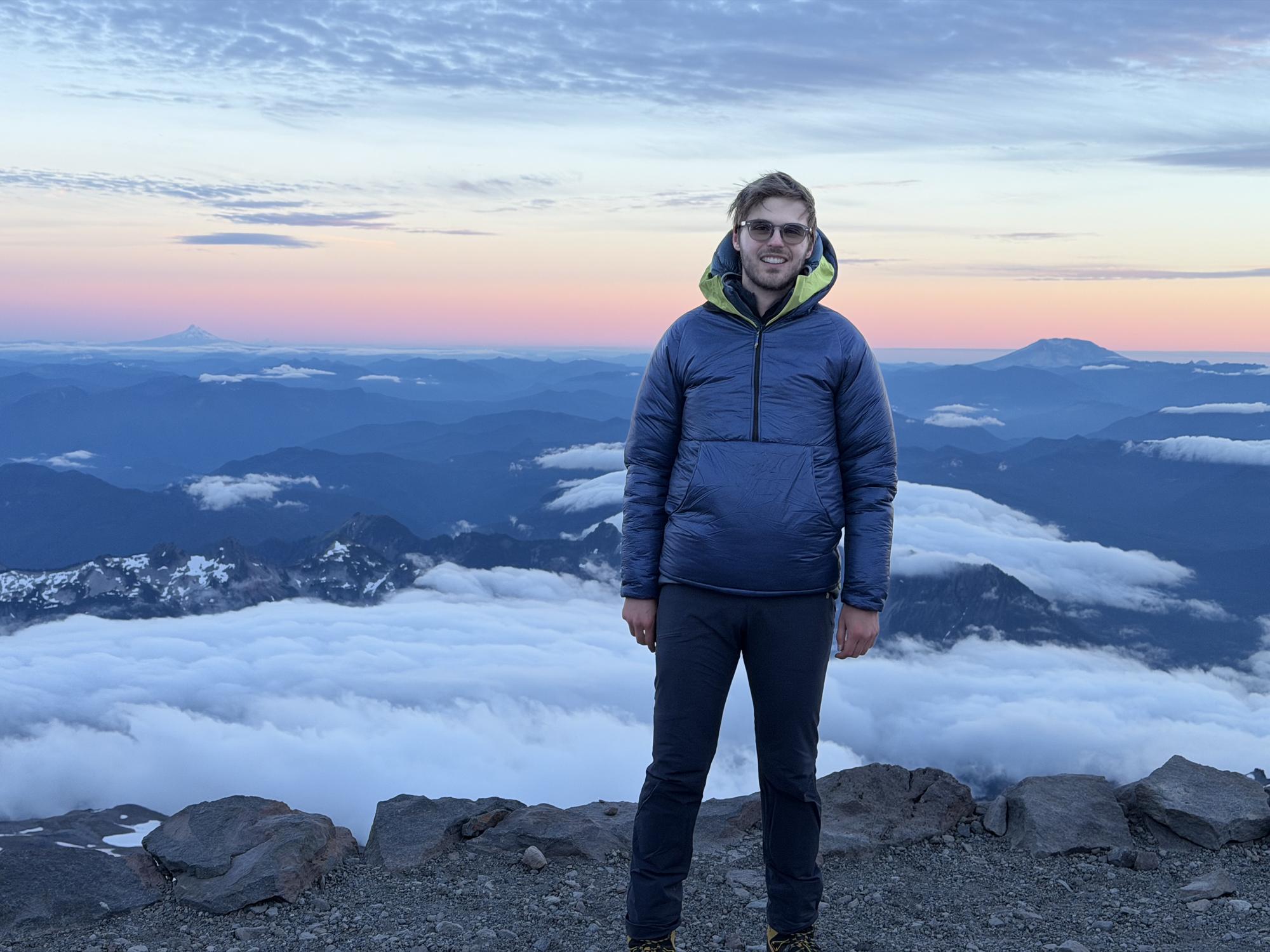 Standing above a sea of clouds at dusk with distant volcanic peaks catching the last light